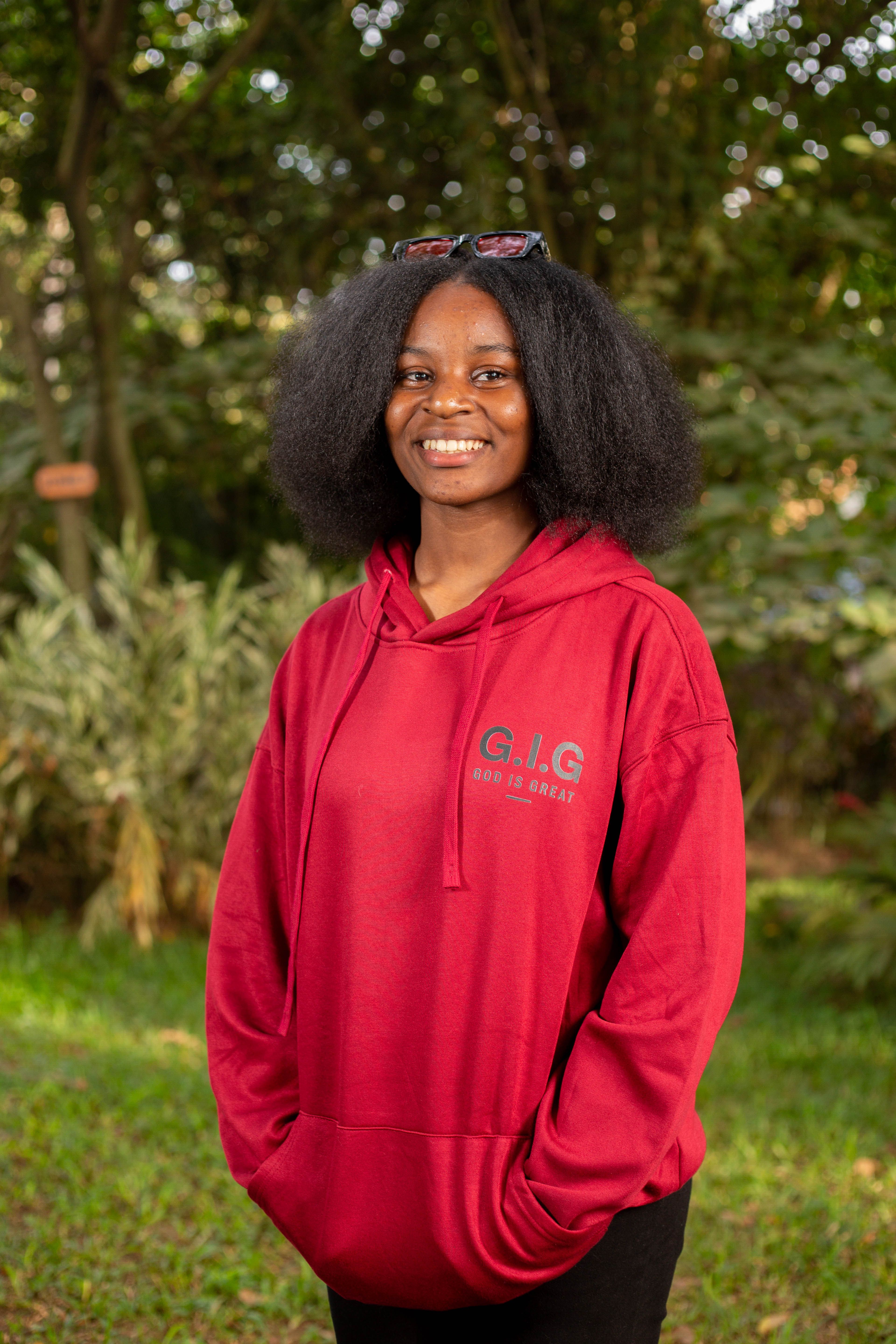 Portrait of a young woman smiling warmly while wearing a red hoodie in a lush outdoor setting.