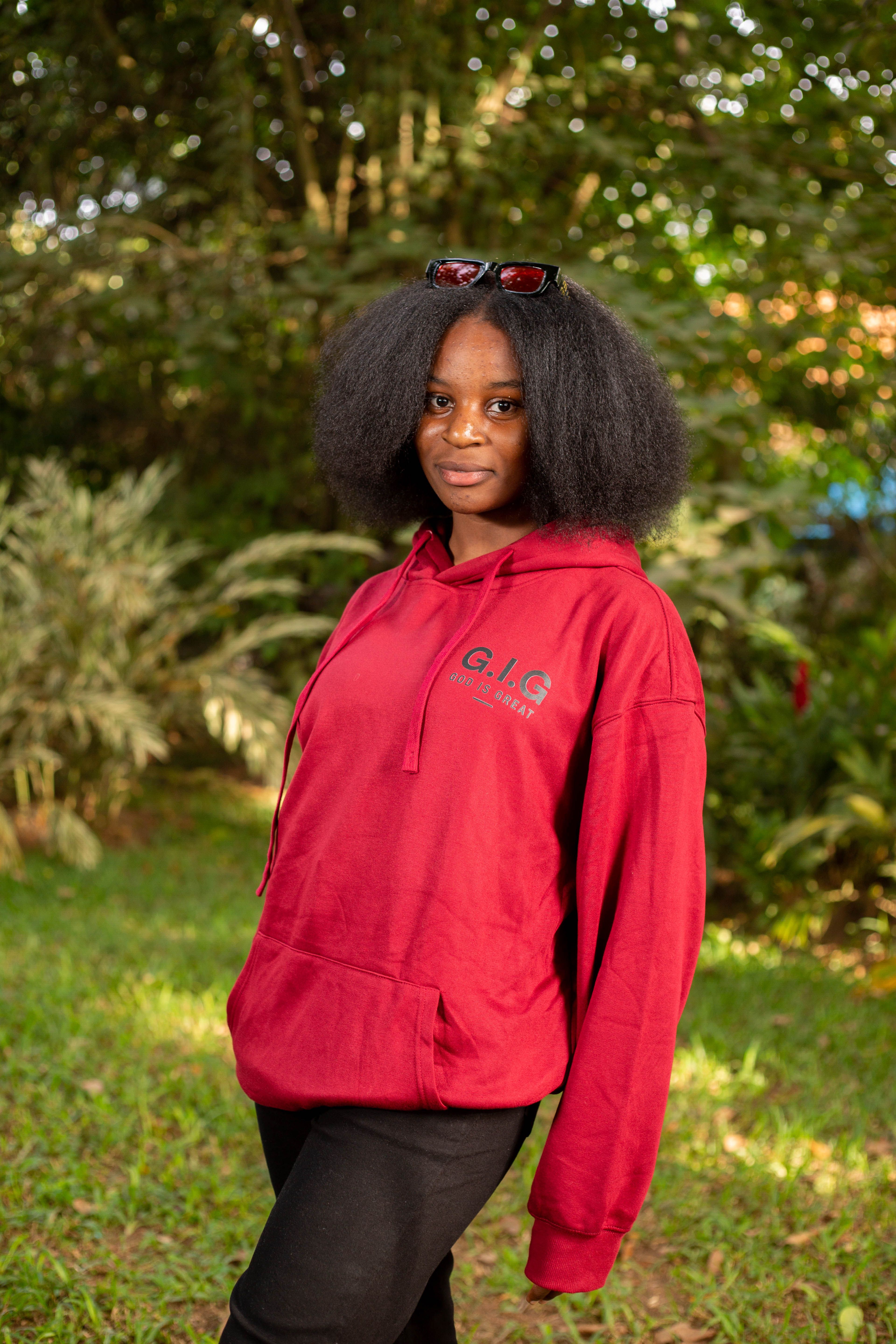 Confident woman with afro hair wearing a red hoodie stands outdoors against a lush green backdrop.