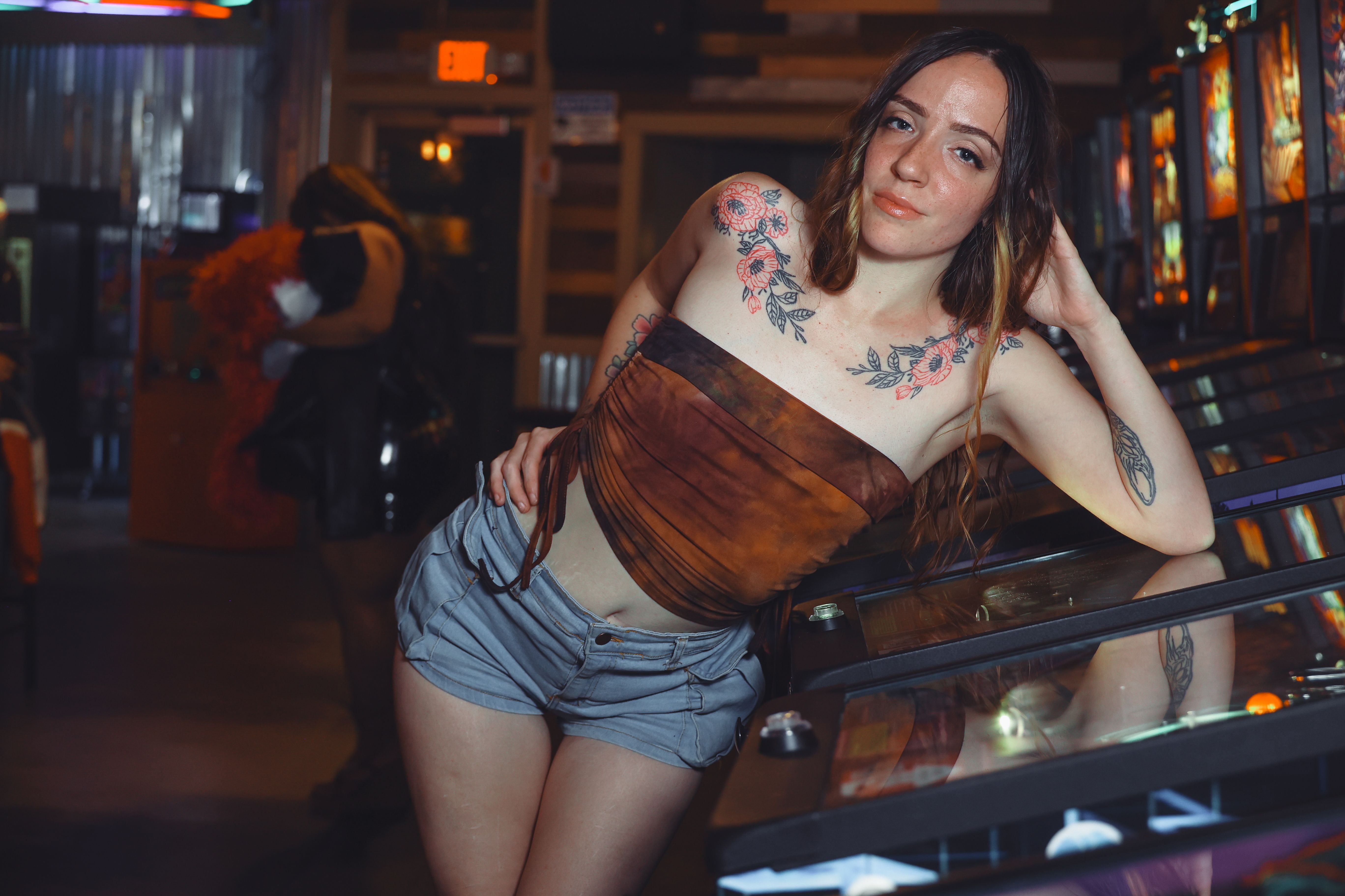 Tattooed woman in casual attire leaning on a pinball machine inside an arcade.