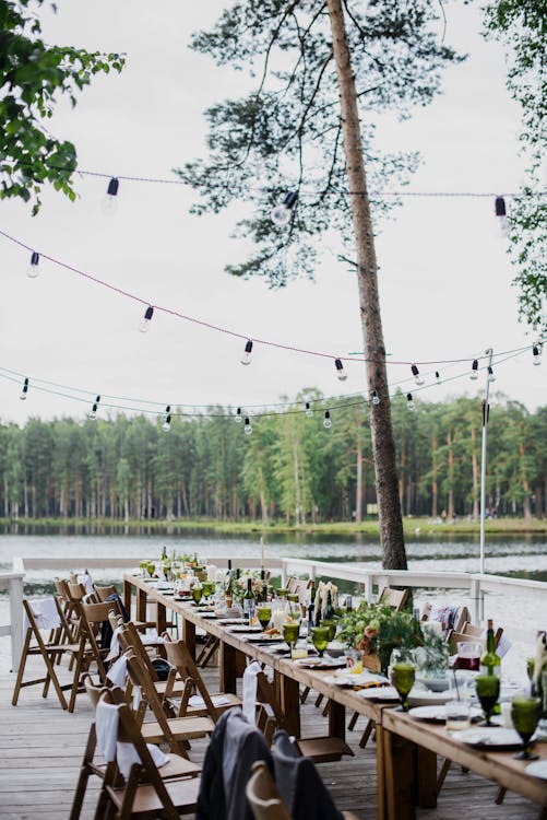Wooden terrace with green trees and tables located near lake preparing for festive romantic party in nature under blue sky