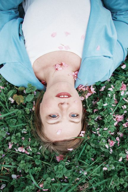 Upside down portrait of a woman lying on grass with pink petals, smiling.