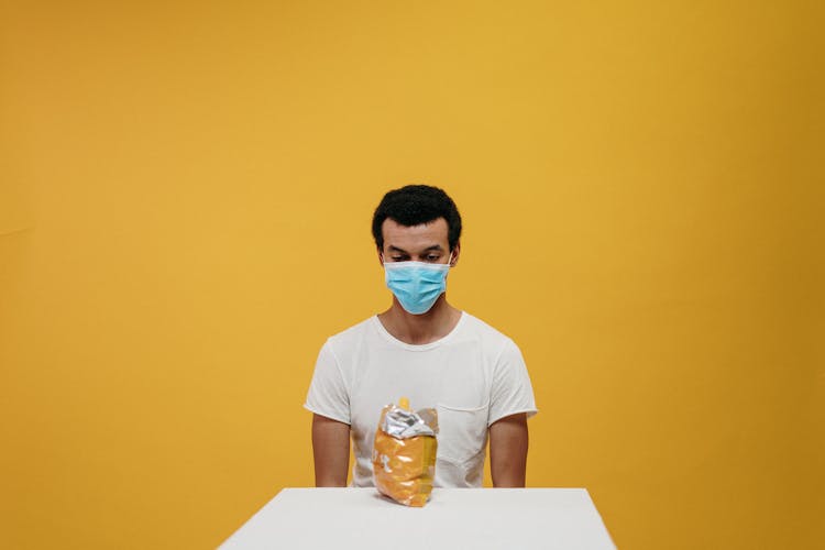 Man In White Crew Neck T-shirt Wearing Blue Face Mask Sitting By The Table