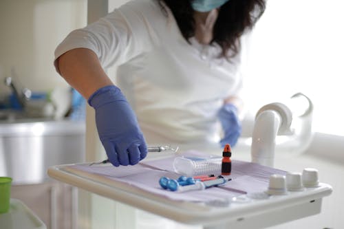 Faceless female stomatologist in blue rubber medical gloves taking instrument from easel in light dental surgery