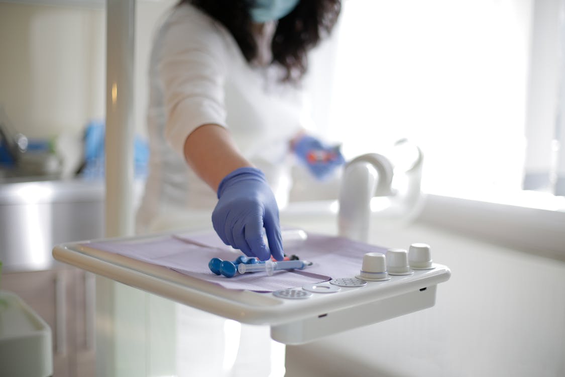 Free Crop unrecognizable female doctor in latex gloves putting syringe and metal pieces of medical tools on dentist table while standing near window in clinic in sunlight Stock Photo