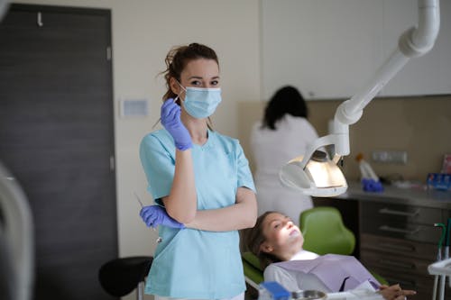 Female assistant and orthodontist in sterile mask and latex gloves taking dental mirror and tweezers while standing near lying female patient and lamp and looking at camera