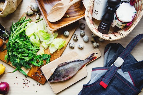 Top view of fresh fish and vegetables put on cutting board near wooden dishware and wicker basket with pepper shaker and jar in kitchen