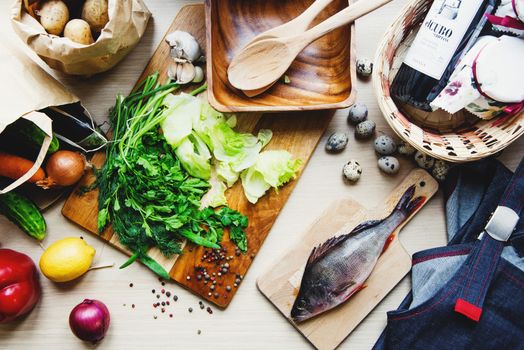 Top view of raw fish put on cutting board near fresh vegetables and wicker basket with bottle of wine and apron at home