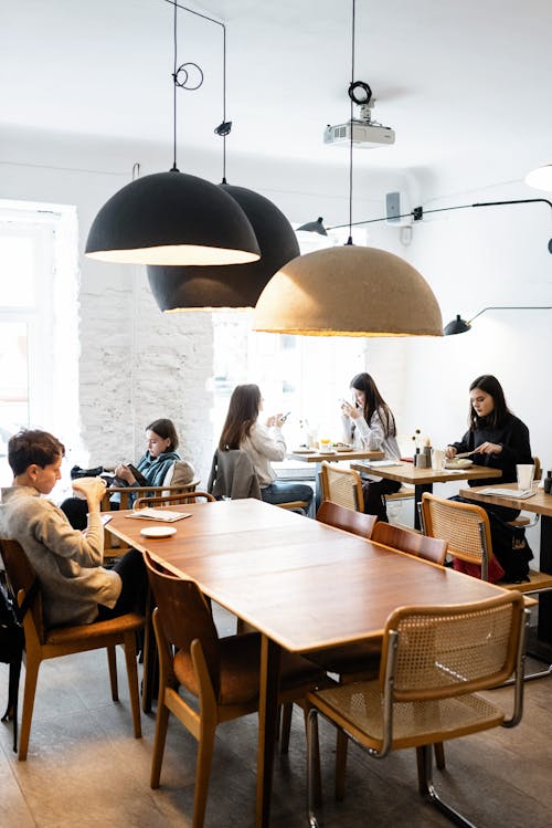 Free Side view of group of female friends talking during lunch time and teenagers sitting at wooden table in cafe with big hanging lamps in daylight Stock Photo