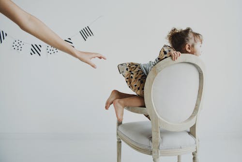Free Side view of crop faceless person reaching out hand to cheerful little female child in dress sitting on classic chair in light studio with garland on wall and looking away Stock Photo