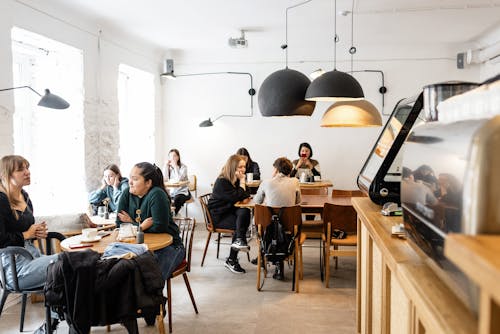 Free Group of multiracial friends in casual wear sitting at wooden tables in cozy cafeteria near cash box and big lamps while conversing Stock Photo