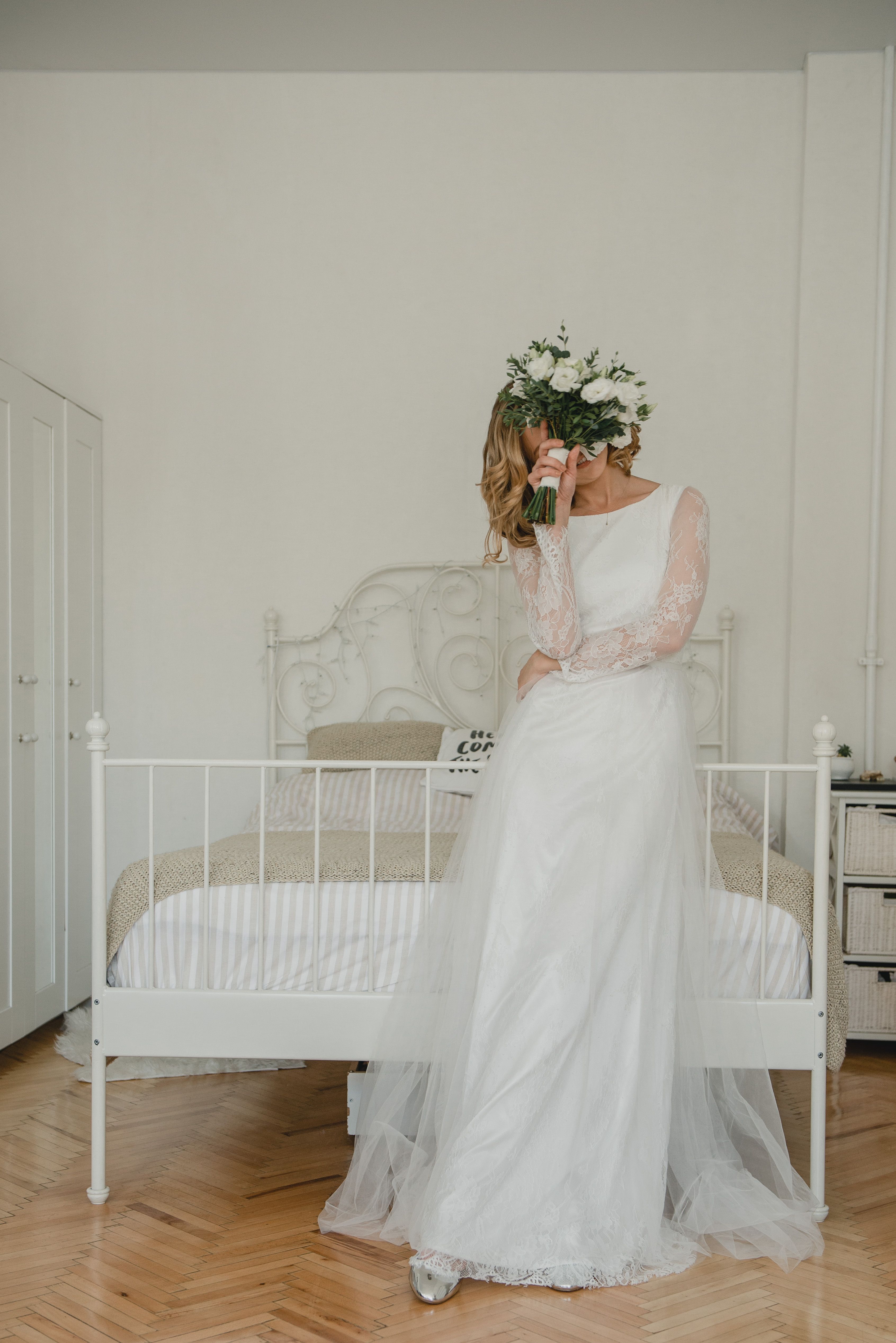 A bride in a white wedding gown holds a bouquet in a serene bedroom setting.