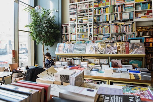 Young female client in casual wear chatting on cellphone while sitting at table with cup of hot drink in cozy bookstore in afternoon