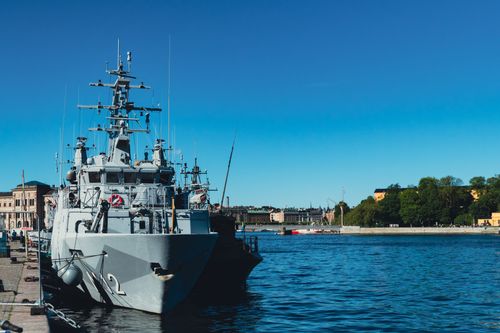 Free A military warship docked in a sunny harbor with clear blue skies and city skyline in the background. Stock Photo