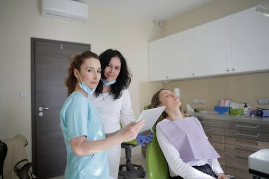 Smiling dentist in sterile mask and uniform looking at camera and showing tablet screen to cheerful female assistant while standing near patient with closed eyes sitting on chair in clinic
