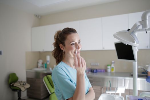 Cheerful young female orthodontist in uniform taking medical tweezers while standing near computer monitor and lamp in clinic and looking away