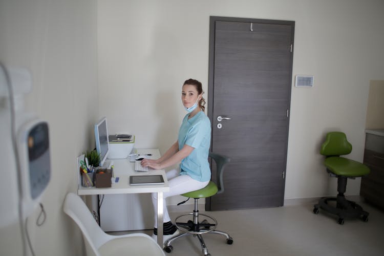 Female Doctor Using Desktop Computer Sitting On Chair In Clinic