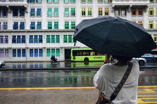 A person with an umbrella watches traffic on a rainy street in Singapore, near vibrant, colorful buildings.