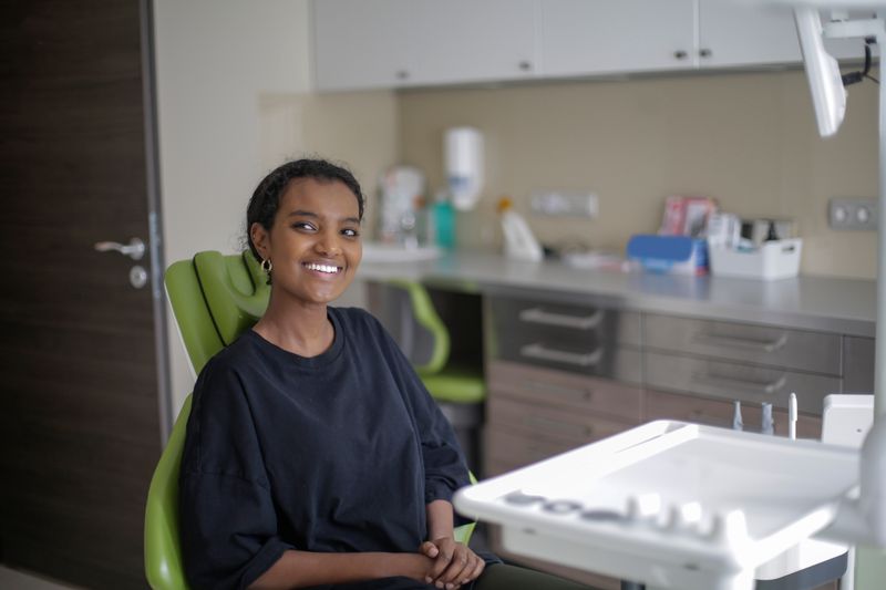 A cheerful young Black woman sits in the exam chair at a dental clinic, with an instrument tray and cabinets behind her. Dental care is one of the services community health centers provide on a sliding fee scale.