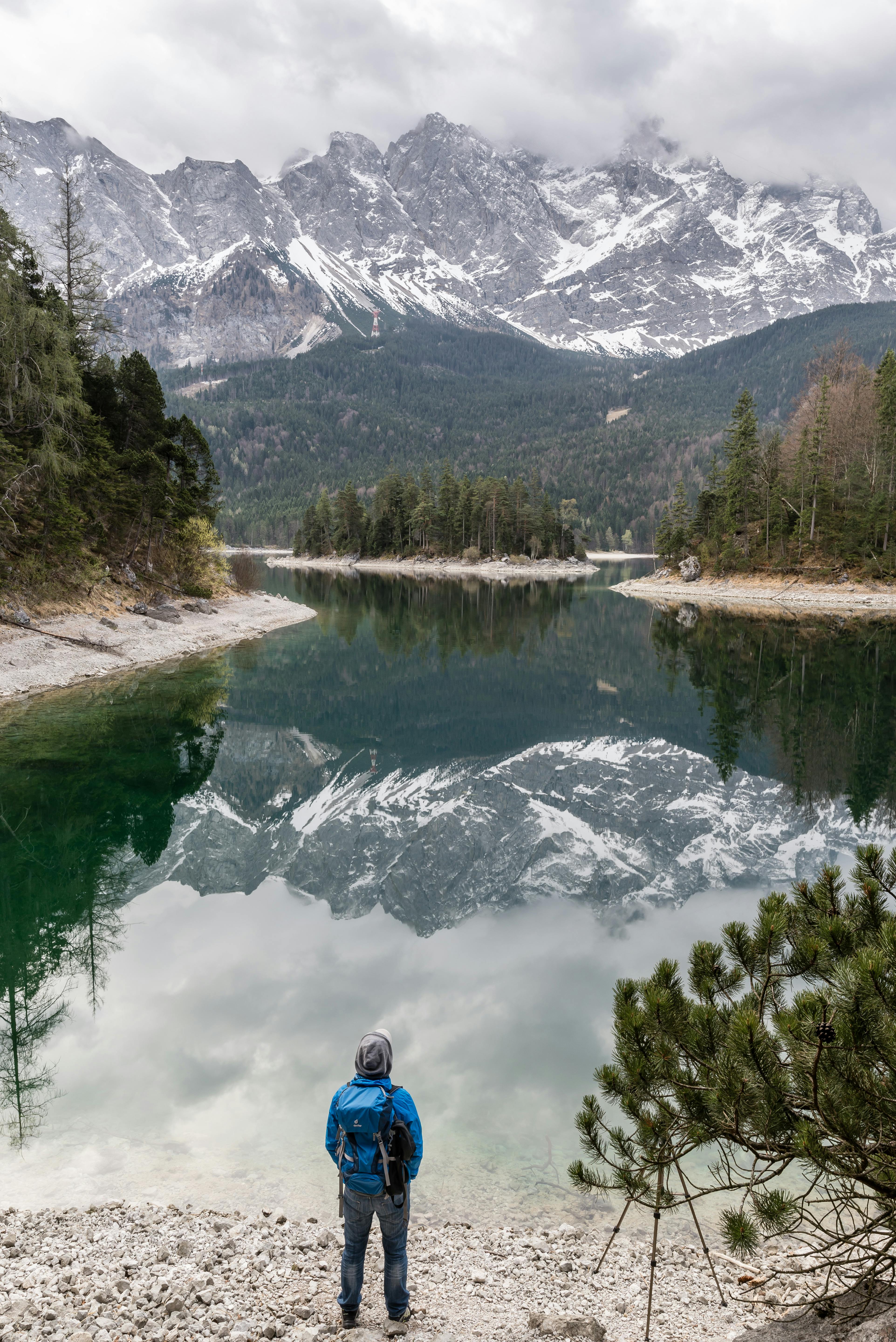 一名男子站在宁静的巴伐利亚湖畔，雪山的优美倒影映在湖中
