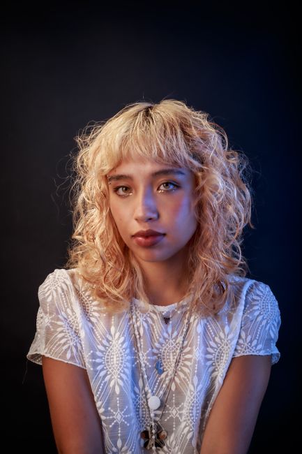 Fotomural Elegante retrato de estudio de una joven con cabello rubio rizado que lleva una blusa blanca de encaje sobre un fondo oscuro.