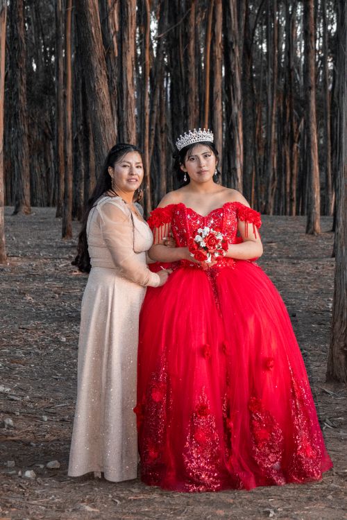 Free Young woman in red quinceañera dress poses with companion among forest trees. Stock Photo