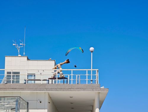 Free Paraglider flying above a rooftop with a clear blue sky. Modern architecture contrasts with flight. Stock Photo