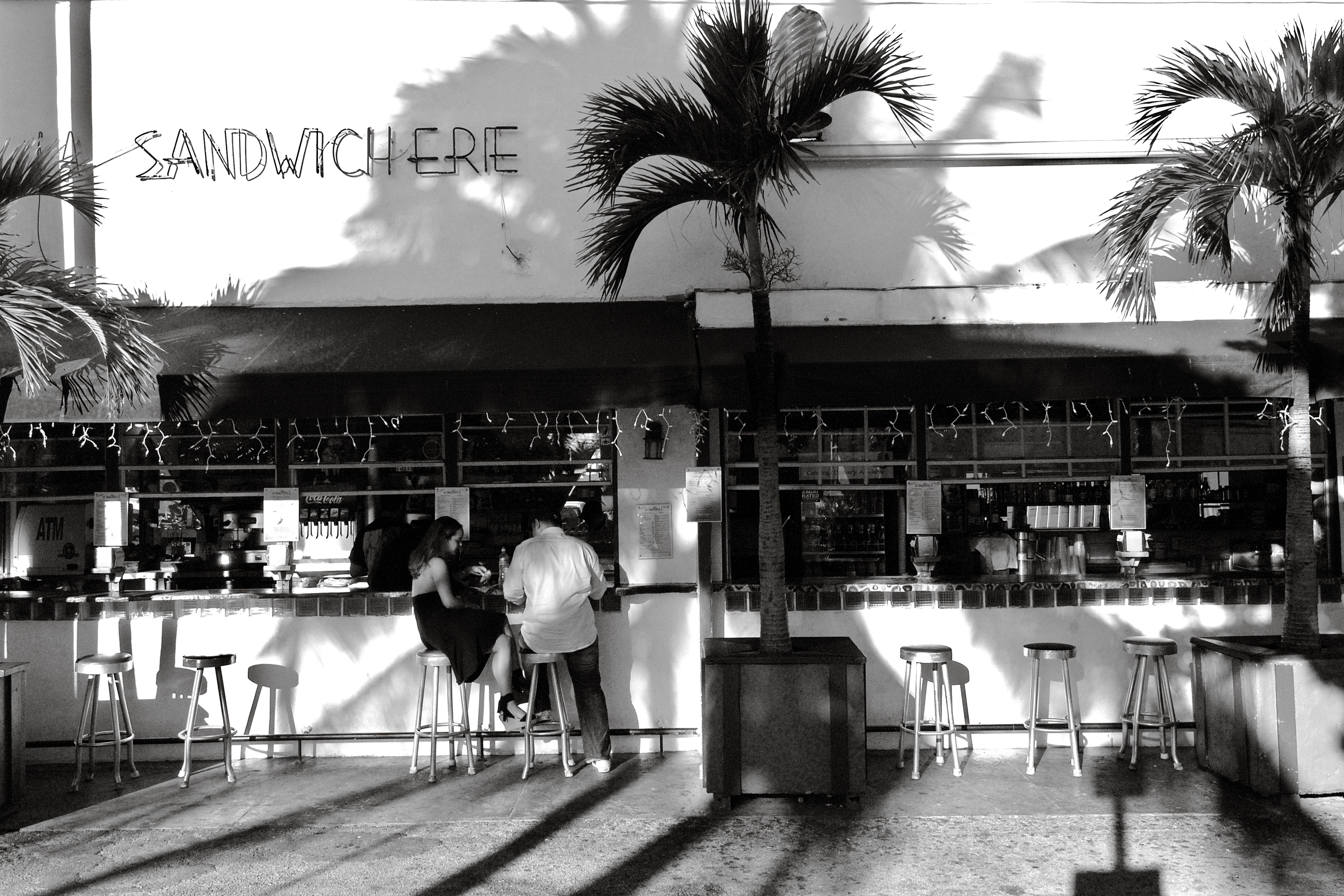 Charming Miami Beach sidewalk sandwich bar with palm tree shadows and patrons enjoying the day.
