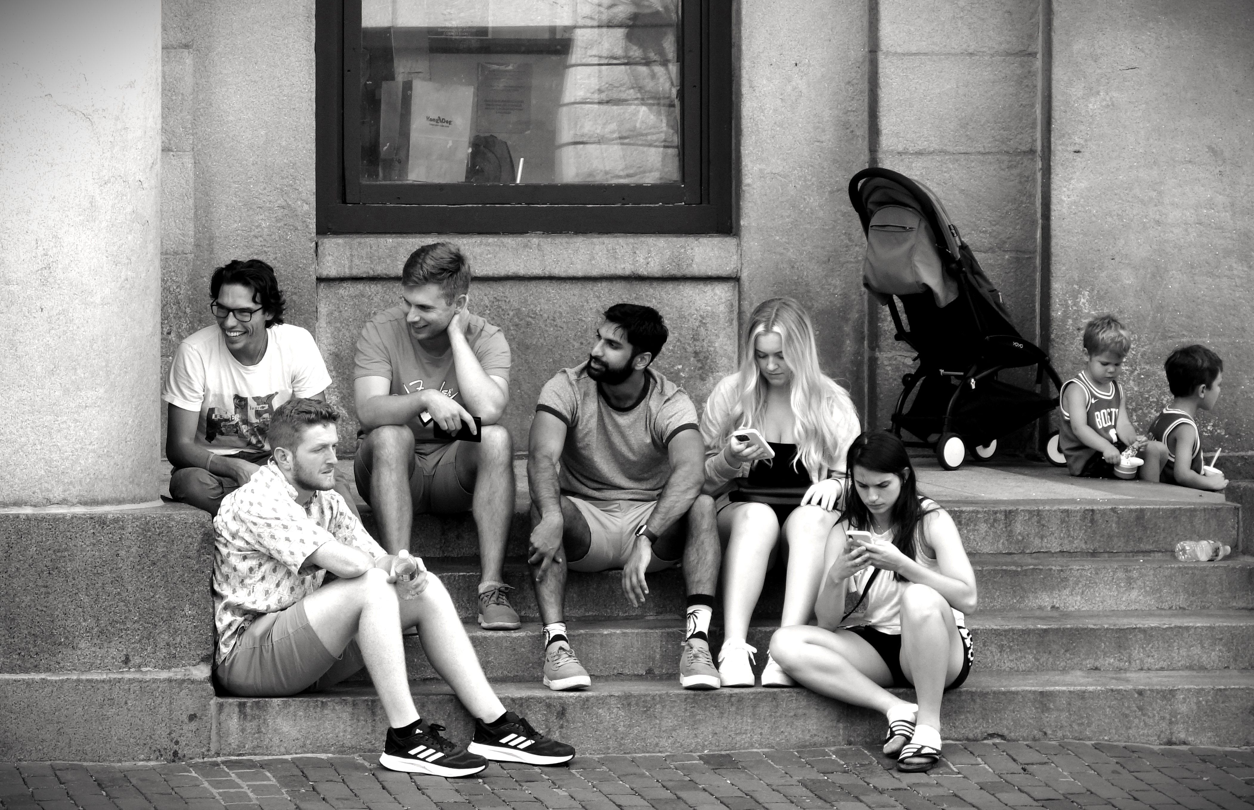 A group of young adults sitting on stone steps, chatting and checking phones, with a stroller nearby.