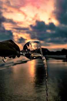 Dramatic silhouette of a hand pouring water from a glass at the beach during sunset.