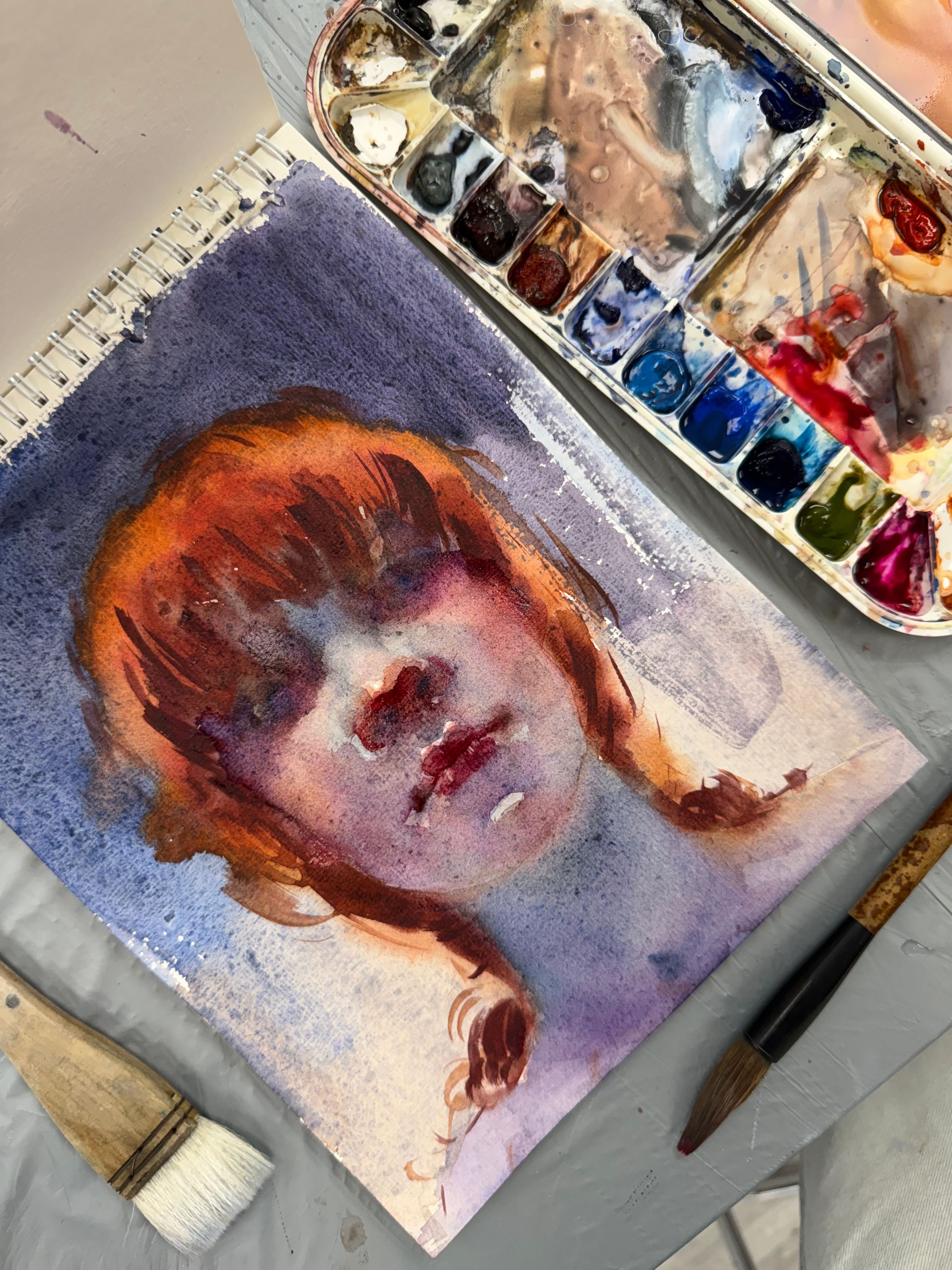 Watercolor painting of a woman with red hair beside a vibrant palette. Artist's tools visible.