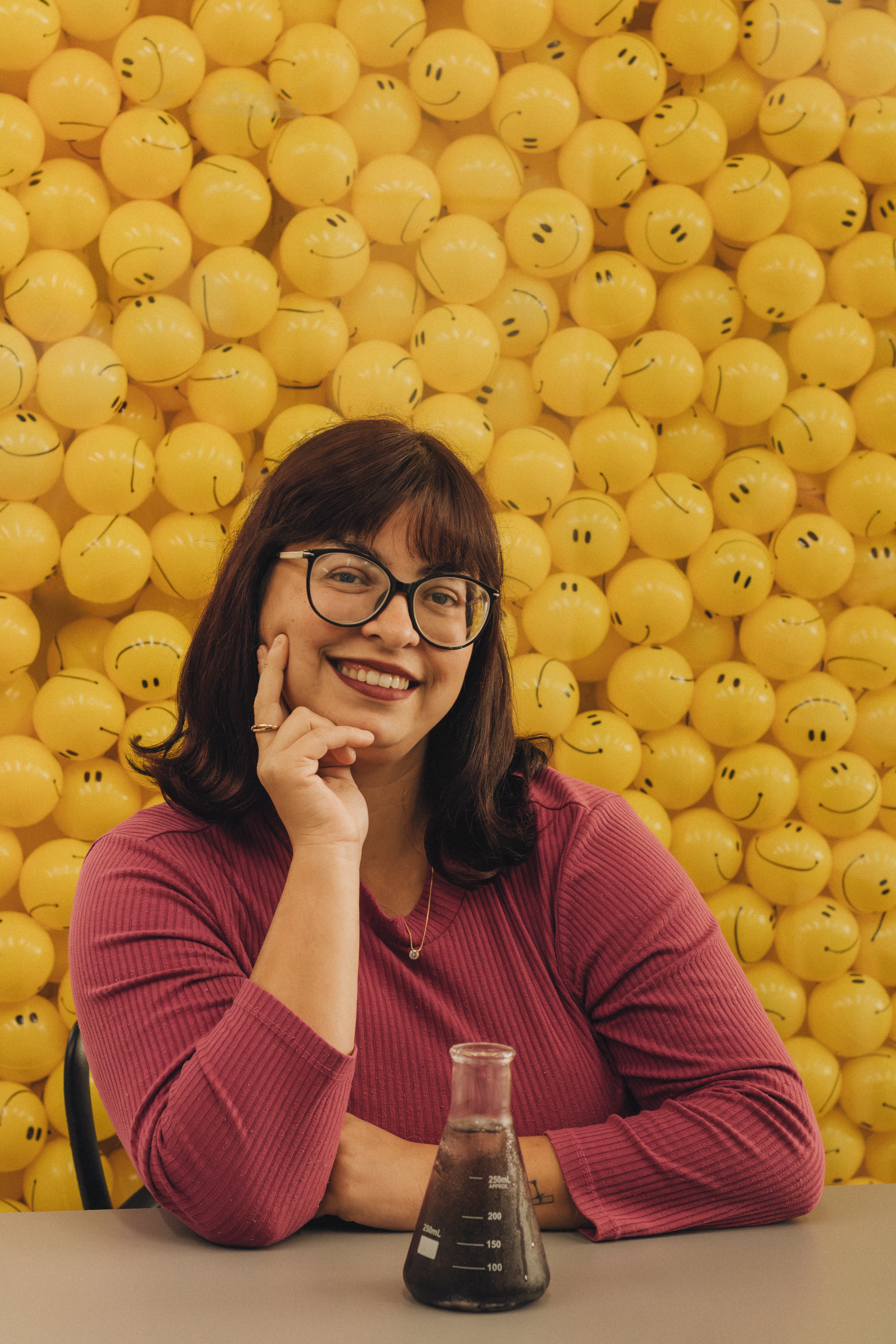Smiling woman in lab with beaker, surrounded by yellow emoji wall.