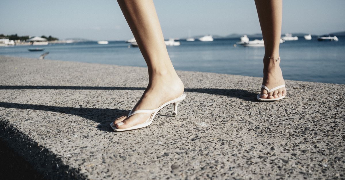 A fashionable close-up of legs in white sandals walking along a seaside promenade on a sunny day.