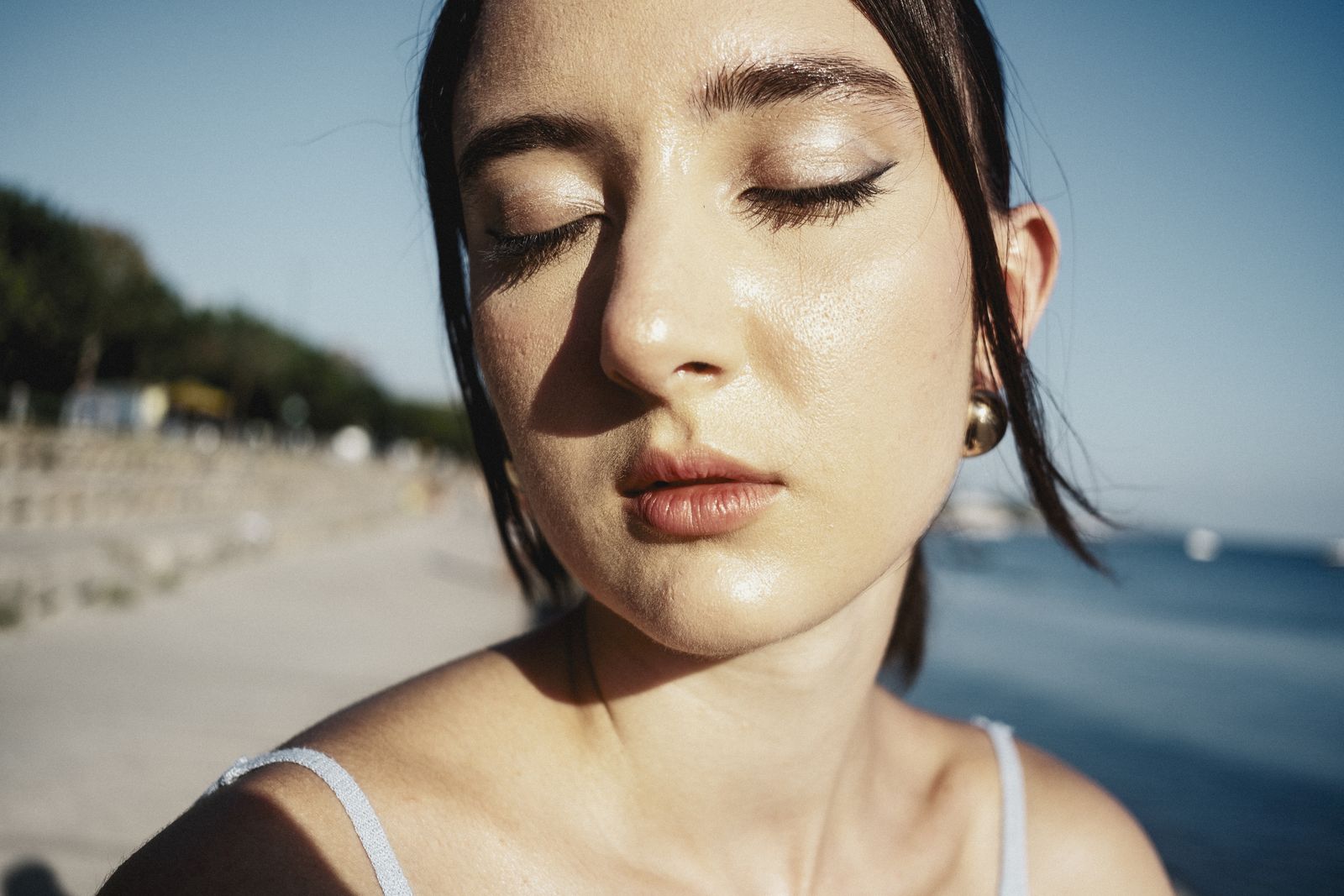Sunlit close-up of a woman with closed eyes and glowing skin against a soft coastal background