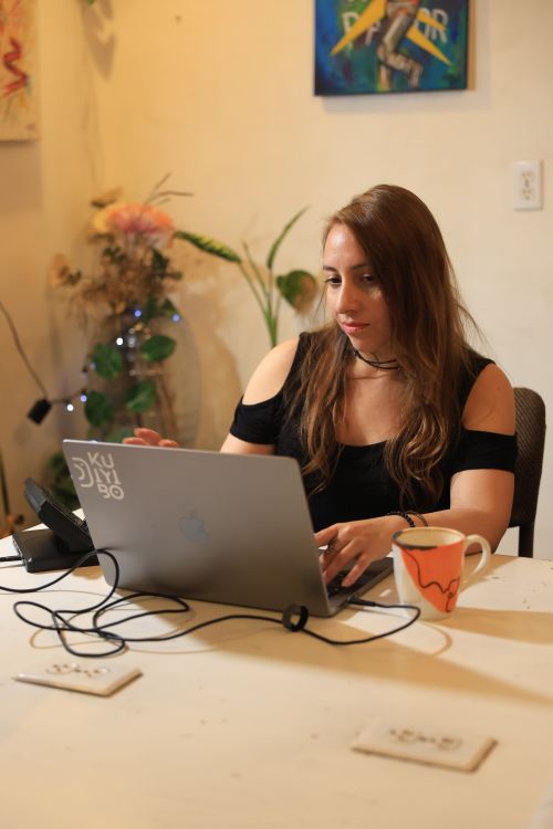 Free Woman working on a laptop at a desk in a warmly lit home office setup with artistic decor. Stock Photo