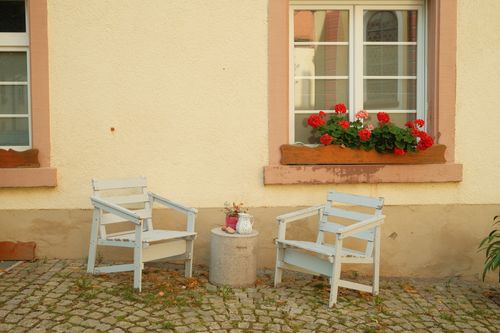 Free Rustic garden chairs and vibrant red flowers under a window create a cozy outdoor setting in Germany. Stock Photo