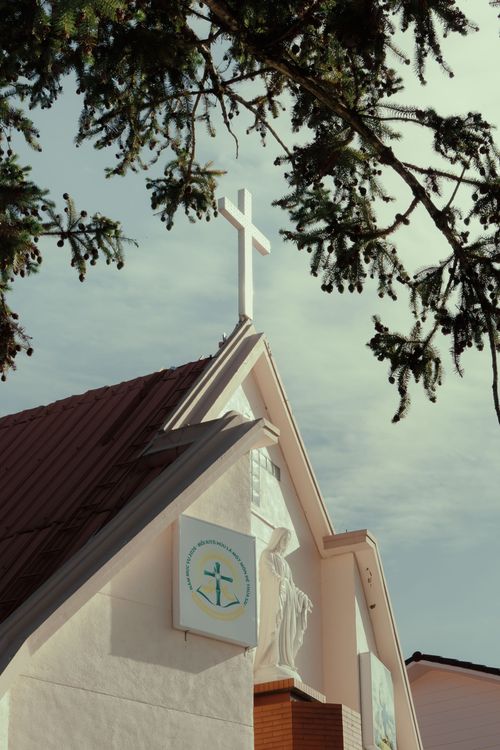 Free A serene view of a church facade with a cross under a clear sky, framed by tree branches. Stock Photo