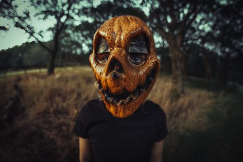 Free A person wearing a pumpkin-themed scary mask stands amidst a dark forest, evoking a Halloween atmosphere. Stock Photo