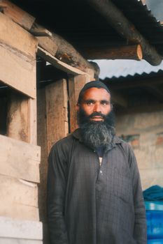 A portrait of a bearded man standing outdoors beside a rustic wooden structure.
