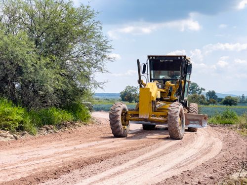 Free A yellow grader smoothing a rural dirt road surrounded by trees and fields under a bright blue sky. Stock Photo
