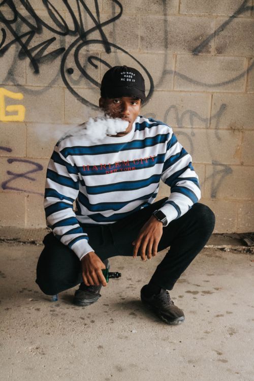 Free Young man crouching while vaping, set against a graffiti-covered wall. Captures urban street style. Stock Photo