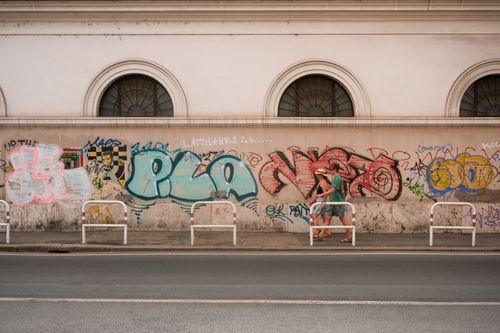 Free Urban scene of two adults walking past a graffiti-covered wall with barred windows in a city. Stock Photo