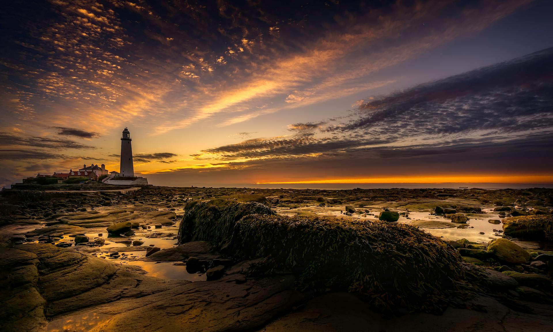 Beautiful lighthouse on rocky coast at sunset with vibrant sky and calm sea reflection.