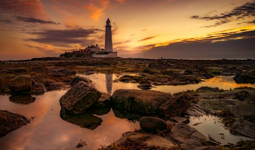 Free Captivating view of a lighthouse at sunset reflected on rocky seashore, creating a serene and dramatic landscape. Stock Photo