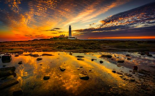 Free A dramatic lighthouse silhouette against a vivid sunset sky, reflecting over tide pools on a rocky coast. Stock Photo