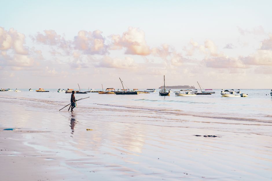 A peaceful beach with a lone person walking and boats on the horizon at sunrise.