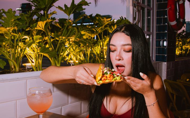 Young woman savoring a taco with a drink at a rooftop restaurant during twilight.