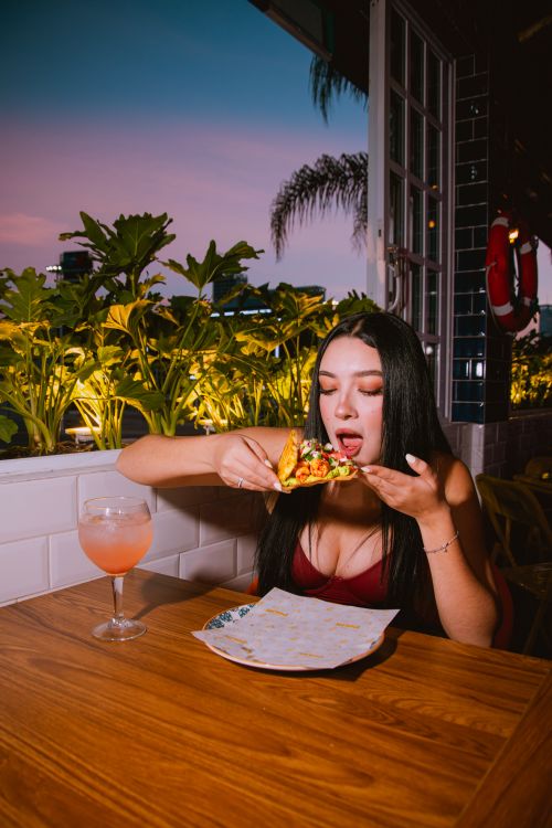 Free Young woman savoring a taco with a drink at a rooftop restaurant during twilight. Stock Photo