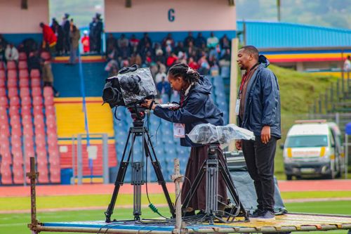 Free Camera operators filming at a stadium in Siteki, Eswatini, capturing a live event. Colorful seating in the background. Stock Photo