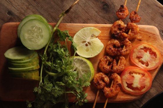 Spiced grilled shrimp skewers on a wooden board with cucumber, lime halves, tomato slices and cilantro