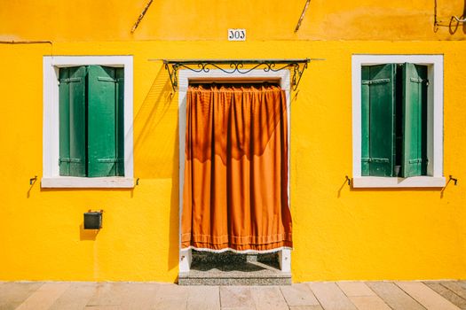 Colorful yellow house exterior with green shutters and an orange curtain door in Burano, Italy.